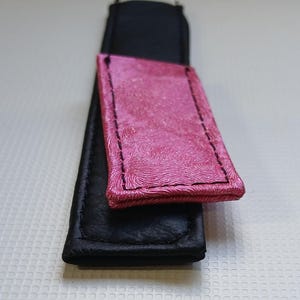 May include: A rectangular black strap featuring a smaller, textured pink rectangular overlay attached to the top. The pink section has a distinct swirling pattern and is secured with black stitching along the edges, contrasting with the smooth black base.