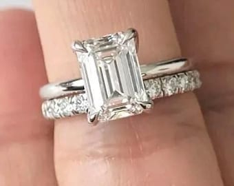 May include: A two-piece ring set featuring a large emerald-cut diamond engagement ring and a delicate diamond band. The rings are set in a silver metal, showcasing the brilliance of the diamonds. The rings are displayed on a light background.