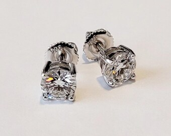 May include: A pair of silver stud earrings featuring round-cut gemstones. The earrings have a post and screw-back fastening. The gemstones are clear and reflect light, creating a brilliant sparkle. The earrings are set on a white surface. The metal is stamped with '925'.