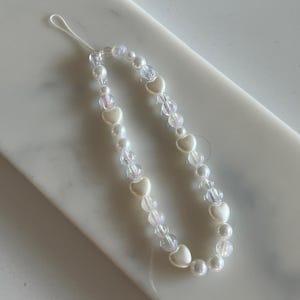 May include: A white beaded phone charm with heart-shaped and clear iridescent beads. The charm is strung on a white cord, creating a delicate accessory. The beads are arranged in a symmetrical pattern. The charm is approximately 15 cm in length.