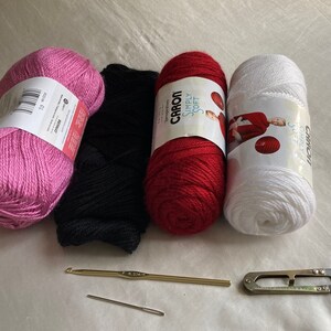 Puede incluir: Cuatro ovillos de lana en rosa, negro, rojo y blanco, acompa&ntilde;ados de un ganchillo dorado, una aguja de coser de metal y unas tijeras de hilo de metal. Las etiquetas del hilo muestran las marcas Bernat y Caron Simply Soft.