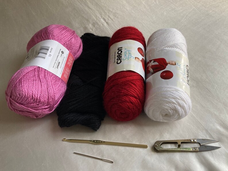 Puede incluir: Cuatro ovillos de lana en rosa, negro, rojo y blanco, acompa&ntilde;ados de un ganchillo dorado, una aguja de coser de metal y unas tijeras de hilo de metal. Las etiquetas del hilo muestran las marcas Bernat y Caron Simply Soft.