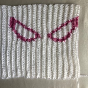 Puede incluir: Una pieza de tela tejida a ganchillo en color blanco con textura acanalada y dos motivos sim&eacute;tricos en forma de ojos angulares delineados con hilo rosa oscuro. El tejido muestra un patr&oacute;n de puntos verticales en toda la superficie.