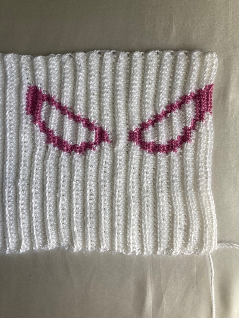 Puede incluir: Una pieza de tela tejida a ganchillo en color blanco con textura acanalada y dos motivos sim&eacute;tricos en forma de ojos angulares delineados con hilo rosa oscuro. El tejido muestra un patr&oacute;n de puntos verticales en toda la superficie.