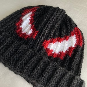 Puede incluir: Un gorro negro tejido con un dise&ntilde;o rojo y blanco. El gorro presenta una textura acanalada y un dise&ntilde;o que se asemeja a la cara de un personaje. El dise&ntilde;o est&aacute; delineado en rojo con relleno blanco.