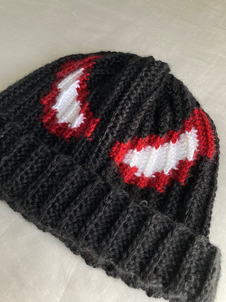 Puede incluir: Un gorro negro tejido con un dise&ntilde;o rojo y blanco. El gorro presenta una textura acanalada y un dise&ntilde;o que se asemeja a la cara de un personaje. El dise&ntilde;o est&aacute; delineado en rojo con relleno blanco.