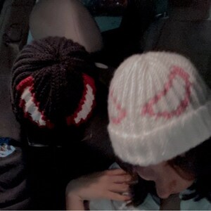 Puede incluir: Dos gorros de punto hechos a mano. Uno es negro con un dise&ntilde;o rojo y blanco, el otro es blanco con un dise&ntilde;o rosa. Los gorros son probablemente para el invierno y est&aacute;n en un coche.