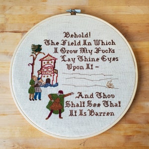 An embroidery hoop with a cross stitched design of 3 people looking at a field with a house and bird in the background. Text reads: Behold! The Field In Which I Grow My Fvcks. Lay Thine Eyes Upon It - And Thou Shalt See That It Is Barren.