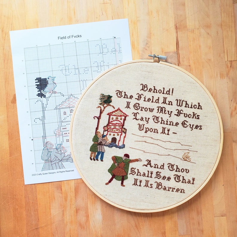 A paper pattern and wooden hoop with a cross stitched design of 3 people looking at a field with a house and bird in the background. Text reads: Behold! The Field In Which I Grow My Fvcks. Lay Thine Eyes Upon It And Thou Shalt See That It Is Barren.