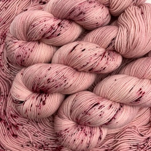 May include: A close-up shot of several skeins of pink yarn with dark red flecks. The yarn is wound into neat, round bundles, with loose strands in the foreground. The colour is a soft, muted pink, with the dark red flecks providing contrast.