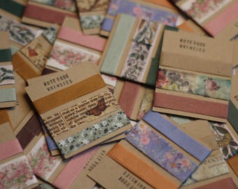 May include: A collection of rectangular kraft paper cards labeled NOTEBOOK WHIMSIES, each wrapped with multiple strips of decorative washi tape featuring floral patterns, vintage newspaper prints, butterfly motifs, and solid colors.