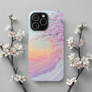 May include: A phone case featuring a pastel landscape design with pink cherry blossoms. The case has a light blue border and a multi-lens camera cutout. The background depicts a mountain range in shades of pink, purple, and orange.
