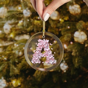 May include: A clear glass ornament with a pink floral design in the shape of the letter A. The ornament is hanging from a gold string.