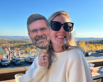 May include: A woman wearing sunglasses and a cream-colored sweater holds a cardboard cutout of a man's face. The background features a scenic overlook with cars and fall foliage under a blue sky.