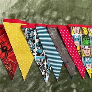 Puede incluir: Una bandera de banderines con siete banderines triangulares rojos, amarillos, azules y grises. Cada banderín presenta un personaje de cómic diferente, incluidos Spider-Man, Capitán América y Hulk. Los banderines están cosidos juntos con hilo rojo.