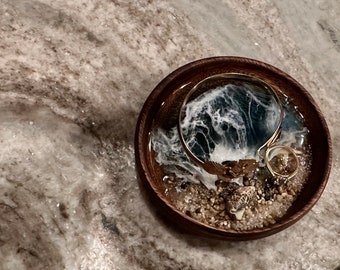 May include: A wooden ring dish featuring a resin ocean scene. The resin is clear and depicts white waves breaking over a dark blue ocean. Gold rings and seashells are placed within the dish.