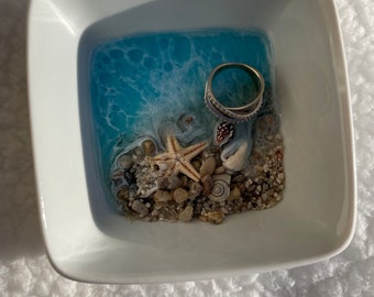 May include: A white ceramic dish with a blue resin ocean scene. The scene includes a starfish, seashells, and small pebbles. A silver ring is resting on the sand.