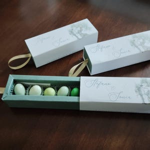 Könnte beinhalten: Drei weiße rechteckige Schachteln mit hellgrünem Innenraum, jede mit einem goldenen Band. Die Schachteln haben eine Baumillustration und die Namen "Stefano & Sonia". Die offene Schachtel enthält fünf bunte Süßigkeiten.