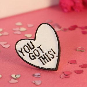 May include: White heart-shaped enamel pin with black outline and the text "You Got This!" on a pink surface with white and pink heart confetti.