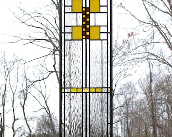 May include: A stained glass window with a geometric design featuring yellow, red, and clear glass panels. The window is framed in black metal and is hanging from a chain.