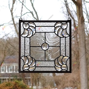 May include: A square stained glass window hanging featuring clear textured glass panels framed in dark metal. The design includes four corner fan-shaped accents and a central circular faceted glass jewel, suspended by two thin metal chains.
