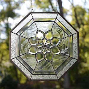 May include: A clear glass octagon-shaped stained glass window with a geometric flower design. The window is hanging from a chain and is in front of a blurred background of green trees.