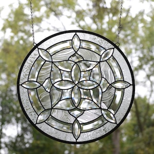 May include: A round stained glass window with a geometric design. The window is made of clear glass with a black metal frame and is hanging from a chain.