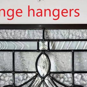 May include: A stained glass window with a geometric design featuring a variety of clear and opaque glass pieces. The window is framed in black metal and has two decorative hangers at the top. The text "1, change hangers" is written above the window.