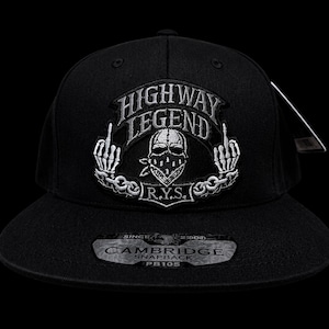 May include: Black snapback hat with the text "HIGHWAY LEGEND" above a skull graphic wearing a bandana. The design includes two hands giving the middle finger. The hat has a flat brim and a tag attached.