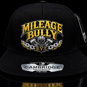May include: Black snapback hat with the embroidered text "MILEAGE BULLY" in yellow, with a skull and engine graphic. The hat also features the text "R.Y.S." and a "CAMBRIDGE SNAPBACK" label. The hat is a PB105 model.
