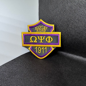 May include: A black and gold Harley Rider patch with purple trim. The patch features the Greek letters "ΩΨΦ" and the year "1911".