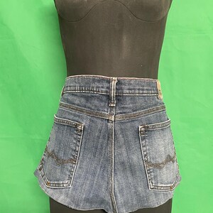May include: Blue denim shorts with two back pockets and decorative stitching. The shorts are displayed on a black mannequin. The shorts have a curved hemline and a faded wash.