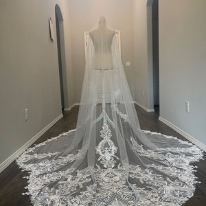 May include: A white bridal veil with a delicate lace pattern. The veil is long and flowing, with a scalloped edge. It is displayed on a mannequin in a hallway.