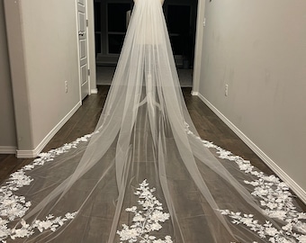 Puede incluir: Un velo de novia largo de tul blanco con un borde de encaje floral. El velo está drapeado sobre un maniquí y se muestra desde atrás.