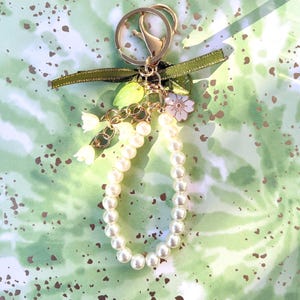 May include: A gold-colored keychain with a pearl beaded loop. The keychain has a green ribbon, small white flower, and leaf charms. The background is a green and white watercolor design with brown speckles.