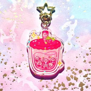 May include: A clear acrylic keychain in the shape of a pink strawberry boba drink. The drink features a pink liquid with small Kirby characters inside. The top has a strawberry and a straw. A gold star-shaped clasp is attached.