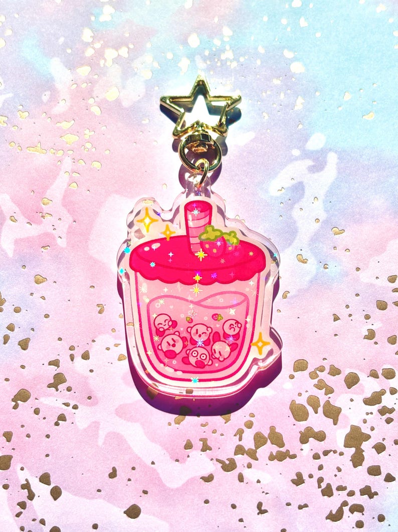 May include: A clear acrylic keychain in the shape of a pink strawberry boba drink. The drink features a pink liquid with small Kirby characters inside. The top has a strawberry and a straw. A gold star-shaped clasp is attached.