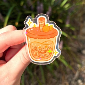 May include: A clear sticker featuring a cartoon-style orange boba tea. The drink has a light orange base with several small, round, cartoon characters inside. The lid is a darker orange with two small orange fruits on top. The sticker has a glittery, holographic border.