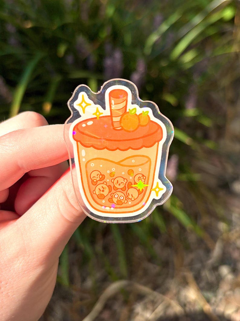 May include: A clear sticker featuring a cartoon-style orange boba tea. The drink has a light orange base with several small, round, cartoon characters inside. The lid is a darker orange with two small orange fruits on top. The sticker has a glittery, holographic border.
