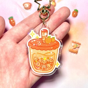 May include: A clear acrylic keychain with a cartoon bubble tea design in orange and yellow. The keychain has a gold star-shaped clasp. Small food stickers are scattered around the keychain. The bubble tea design includes cartoon characters.