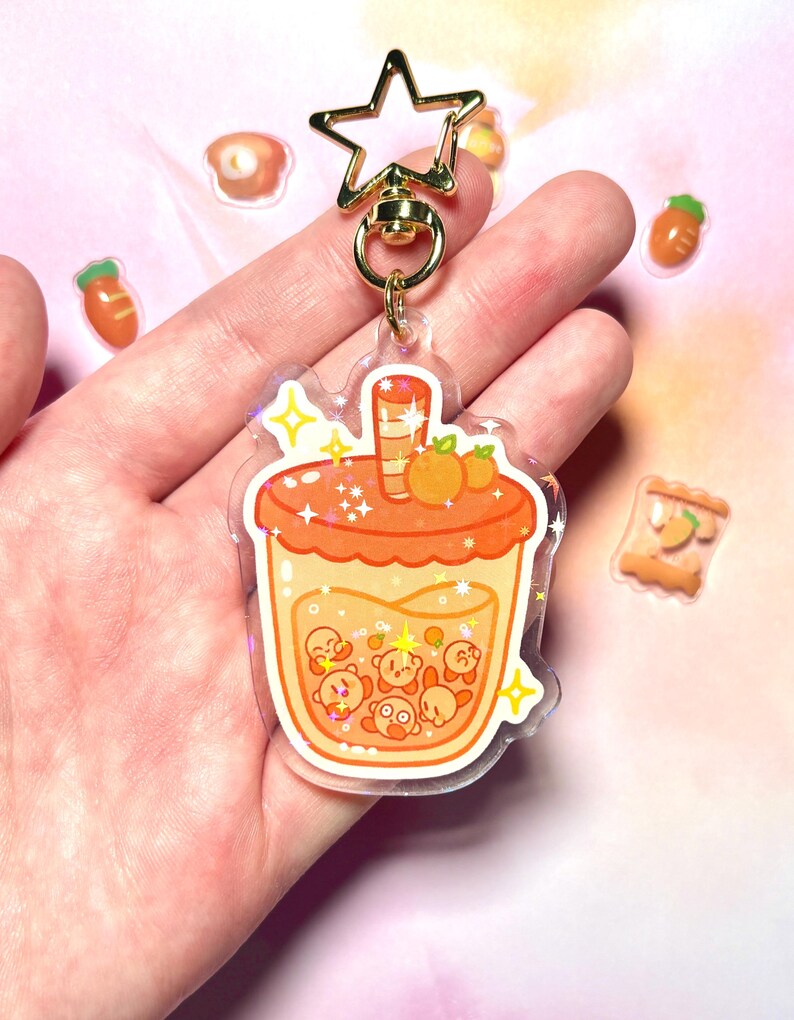May include: A clear acrylic keychain with a cartoon bubble tea design in orange and yellow. The keychain has a gold star-shaped clasp. Small food stickers are scattered around the keychain. The bubble tea design includes cartoon characters.
