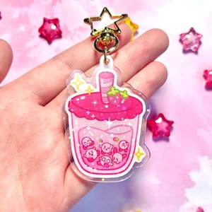 May include: A pink acrylic charm shaped like a bubble tea drink, with a strawberry and cartoon characters. It has a gold star-shaped clasp and ring. The background is light pink with star decorations. The charm is a fun accessory.