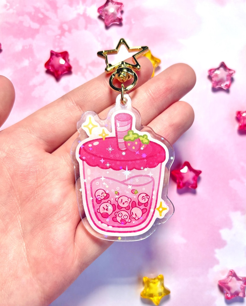 May include: A pink acrylic charm shaped like a bubble tea drink, with a strawberry and cartoon characters. It has a gold star-shaped clasp and ring. The background is light pink with star decorations. The charm is a fun accessory.