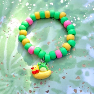 Puede incluir: Un brazalete de cuentas verdes, amarillas y rosas con un dije de una rana montando un pato amarillo.