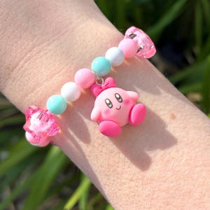 Pu&ograve; includere: Un braccialetto di perline rosa e bianche con un ciondolo rosa e bianco di un personaggio dei cartoni animati. Il ciondolo ha un fiocco rosa e un viso rosa con occhi neri e un sorriso.