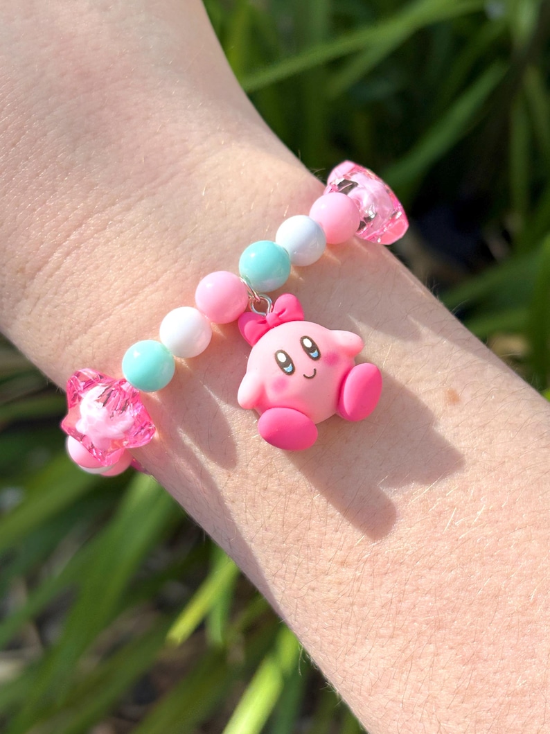 Pu&ograve; includere: Un braccialetto di perline rosa e bianche con un ciondolo rosa e bianco di un personaggio dei cartoni animati. Il ciondolo ha un fiocco rosa e un viso rosa con occhi neri e un sorriso.