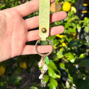 May include: A light green leather keychain with a gold-colored key ring. The keychain has a textured, crocodile-like pattern and a gold-colored rivet. Dangling from the ring are small white flower charms and green leaf charms.