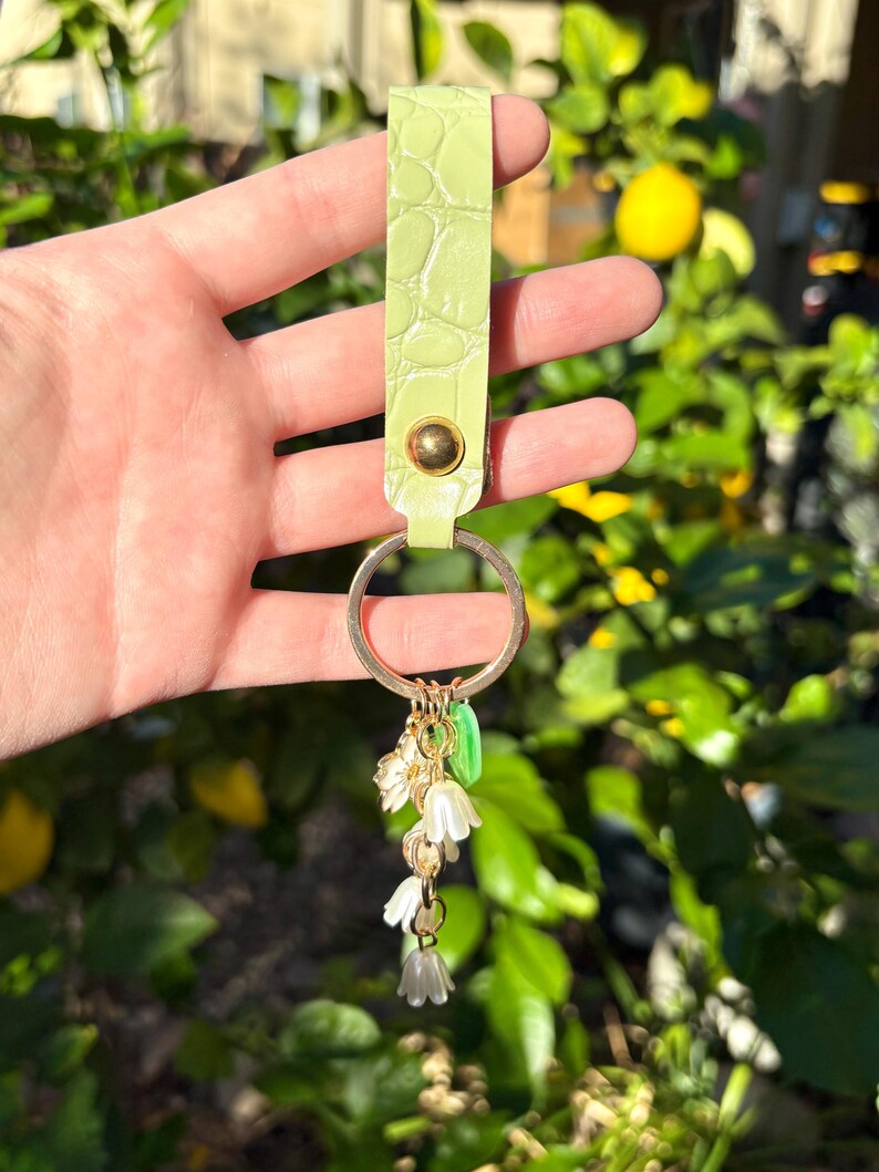 May include: A light green leather keychain with a gold-colored key ring. The keychain has a textured, crocodile-like pattern and a gold-colored rivet. Dangling from the ring are small white flower charms and green leaf charms.
