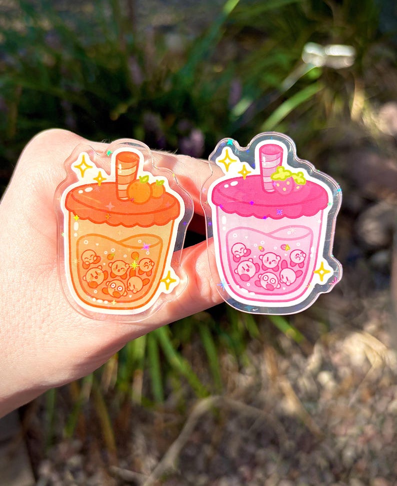 May include: Two clear acrylic bubble tea charms. One is orange with orange fruit and the other is pink with a strawberry. Both charms have a cartoon character inside the drink and are decorated with yellow stars.