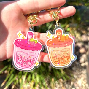 May include: Two acrylic keyrings with bubble tea designs. One is pink with a strawberry, the other is orange with oranges. Both keyrings have gold-coloured clasps and include cartoon characters inside the drinks.